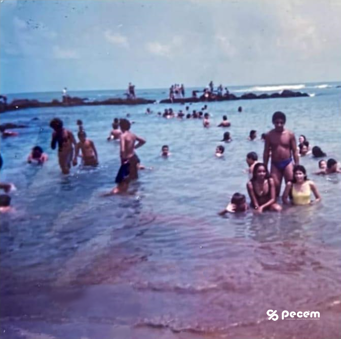 Foto de morador da comunidade com público na praia do Pecém e ao fundo parte da antiga estrutura do primeiro porto, do século XIX.